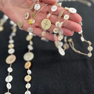 May include: A string of white and cream buttons strung together with twine. The buttons are various sizes and shapes, and some have a floral design.