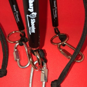 May include: Three black retractable pens with black lanyards and silver carabiners. The pens have the text "Sharp Shooter" printed on them.