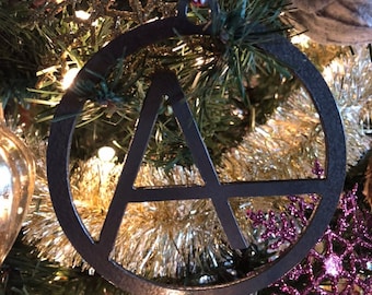 May include: A black metal Christmas bauble shaped like a circle with an 'A' inside. The bauble is hanging on a Christmas tree with gold tinsel and purple glitter snowflakes.