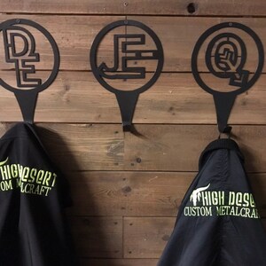 May include: Three black metal wall hooks with circular designs and letter initials, hanging on a wooden wall. Below the hooks are black jackets with the text "HIGH DESERT CUSTOM METALCRAFT" in neon green.