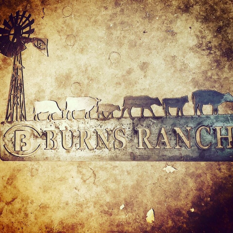 May include: A metal sign with the words "Burns Ranch" in a stylised font. The sign features a silhouette of a windmill and a line of cattle walking across a field.