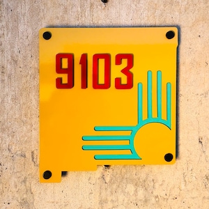 Pode incluir: Um sinal de número de casa de metal amarelo com o número 9103 em vermelho e um símbolo Zia azul turquesa.