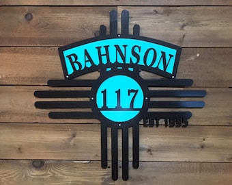 May include: Black metal wall sign with the name "BAHNSON" in white letters on a turquoise background. The sign also features the number "117" and the text "EST 1995". The sign is in the shape of a sun symbol.