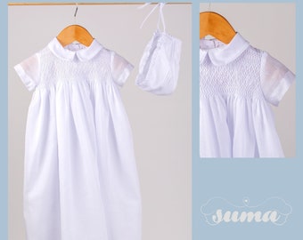 Könnte beinhalten: Weißes Taufkleid mit einem Smocksaum und einer passenden Haube. Das Kleid hat einen Peter-Pan-Kragen und kurze Ärmel. Das Kleid hängt an einem Holzbügel.