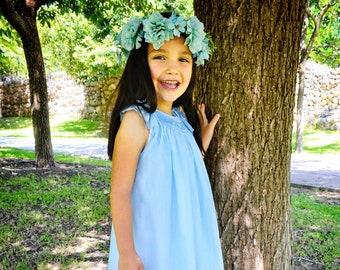 Puede incluir: Una niña pequeña con un vestido azul claro y una corona de flores se para junto a un árbol. Está descalza y sonriendo.