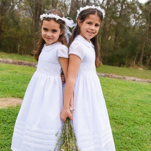 Op de afbeelding: Twee jonge meisjes in witte jurken en bloemenkransen staan ​​naast elkaar in een grasveld. Ze houden een boeket witte bloemen vast.