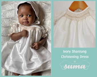 May include: A white christening dress made of ivory shantung fabric. The dress has a smocked bodice and a full skirt. The dress is shown on a baby wearing a white bonnet.