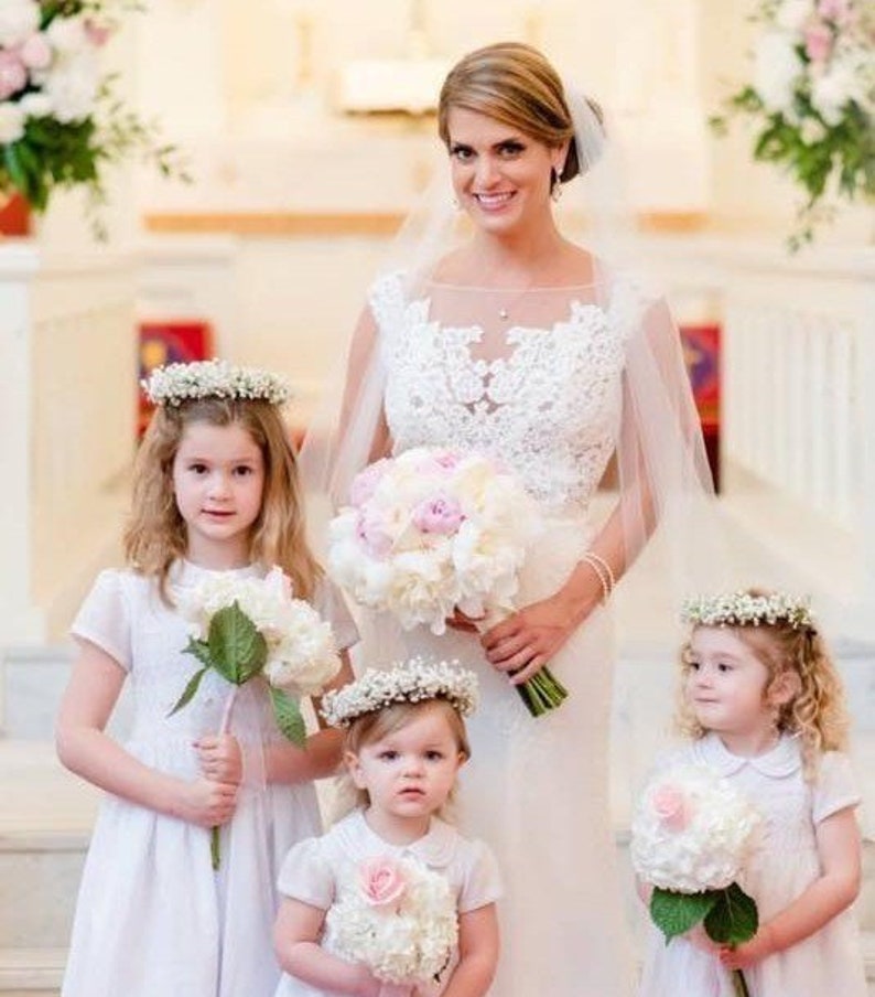 Op de afbeelding: Een bruid in een witte kanten trouwjurk met een sluier houdt een boeket met witte en roze bloemen vast. Drie bruidsmeisjes in witte jurken dragen bloemenkransen en houden boeketten met witte bloemen vast.