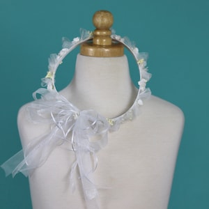 Op de afbeelding: Witte bloemenkrans met een witte tule strik. De krans is gemaakt van wit stof en heeft kleine witte bloemen en groene bladeren.
