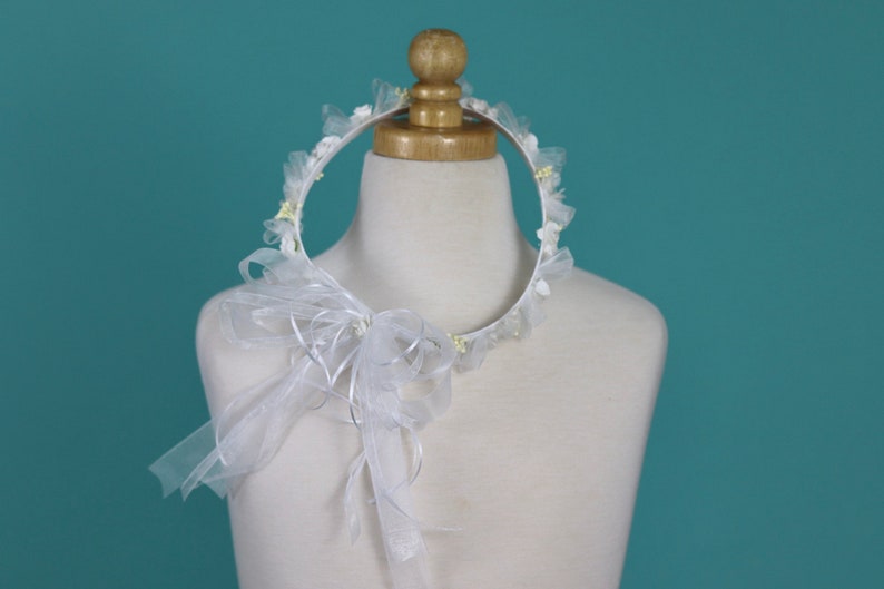 Op de afbeelding: Witte bloemenkrans met een witte tule strik. De krans is gemaakt van wit stof en heeft kleine witte bloemen en groene bladeren.
