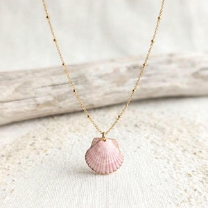 Peut inclure: Un collier à fine chaîne dorée agrémenté d'un petit pendentif en forme de coquille Saint-Jacques rose clair. La chaîne est ornée de perles dorées régulièrement espacées, et le coquillage présente une texture nervurée et une teinte rose naturelle.