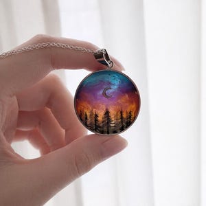 May include: A circular silver pendant necklace featuring a colourful resin inlay of a night sky with a crescent moon above a dark forest silhouette. The sky transitions from deep blue and purple at the top to a warm orange glow near the horizon.