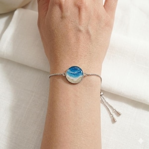 May include: A silver-toned adjustable bracelet featuring a circular charm with a resin inlay depicting a miniature ocean scene. The design includes shades of blue for the water, white for the foam, and a sandy beige colour for the shoreline.