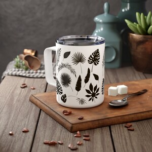 May include: White ceramic mug with a black tropical leaf design. The mug has a lid and a handle.