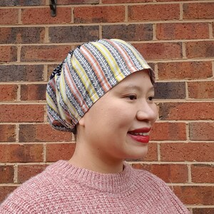 Peut inclure: Un couvre-chef à motifs avec un mélange de rayures noires, blanches, jaunes et oranges. Le couvre-chef est porté par une personne portant un pull rose. L'arrière-plan est un mur de briques.