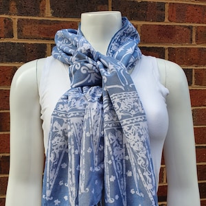 Op de afbeelding: Blauw-witte sjaal met patroon en kwastjes. De sjaal is om de nek van een mannequin gedrapeerd en toont het ingewikkelde ontwerp. Het patroon heeft bloem- en geometrische elementen. De randen van de sjaal zijn afgewerkt met delicate kwastjes.