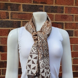 Op de afbeelding: Een bruin en wit patroon sjaal met geometrische en abstracte ontwerpen is om de nek van een mannequin gedrapeerd. De sjaal heeft een donkerbruine rand en kwastjes. De mannequin draagt een witte tanktop.