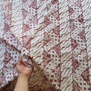 Könnte beinhalten: Cremefarbener Stoff mit einem sich wiederholenden Muster in Burgunderrot. Das Design zeigt stilisierte, geschwungene Linien und geometrische Formen. Der Stoff ist drapiert und zeigt Falten und Textur. Die Gesamtästhetik deutet auf ein traditionelles Textil hin.