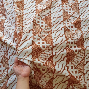 Könnte beinhalten: Cremefarbener Stoff mit braunem Batikmuster. Das Design zeigt sich wiederholende vertikale Bänder aus stilisierten, geschwungenen Formen und geometrischen Elementen. Der Stoff ist gefaltet und zeigt die Textur und den Fall des Materials.