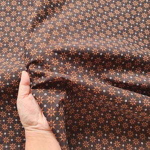 May include: Dark brown fabric featuring a repeating pattern of orange and white starburst designs. The fabric is held up, displaying its texture and drape. The pattern is evenly spaced across the material.
