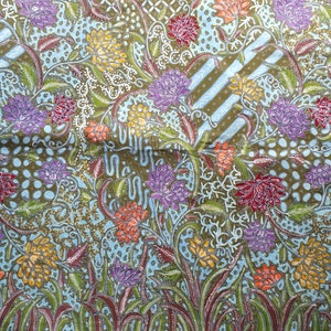 Könnte beinhalten: Ein detaillierter Stoff mit floralem Muster. Das Design zeigt große, stilisierte Blumen in Lila, Gelb, Orange und Rot, vor einem hellblauen und olivgrünen Hintergrund. Der Stoff hat einen Rand mit einem sich wiederholenden Punktmuster.