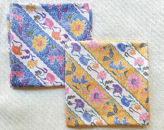 May include: Two folded batik fabric pieces with floral patterns. One features a blue and white design with pink, orange, and yellow flowers. The other has a yellow and white design with pink, purple, and blue flowers. The fabric is folded.