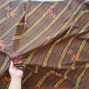 以下が含まれることがあります： 黄色と黒の斜めストライプ模様の茶色の生地。生地には赤、オレンジ、緑の花柄があります。生地は折りたたまれ、手で持たれています。