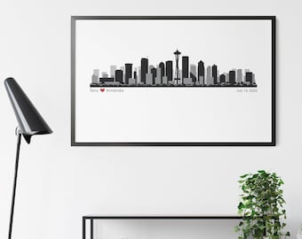 Puede incluir: Silueta en blanco y negro del horizonte de Seattle con la Space Needle, un corazón con los nombres "Tony" y "Amanda" y la fecha "July 14, 2022". La imagen está enmarcada y colgada en una pared blanca sobre un estante de madera con libros y decoración.