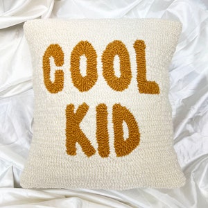 May include: Cream-colored square pillow with the words "COOL KID" embroidered in a textured, golden-brown font. The pillow has a soft, plush texture, and the text is centered on the front.