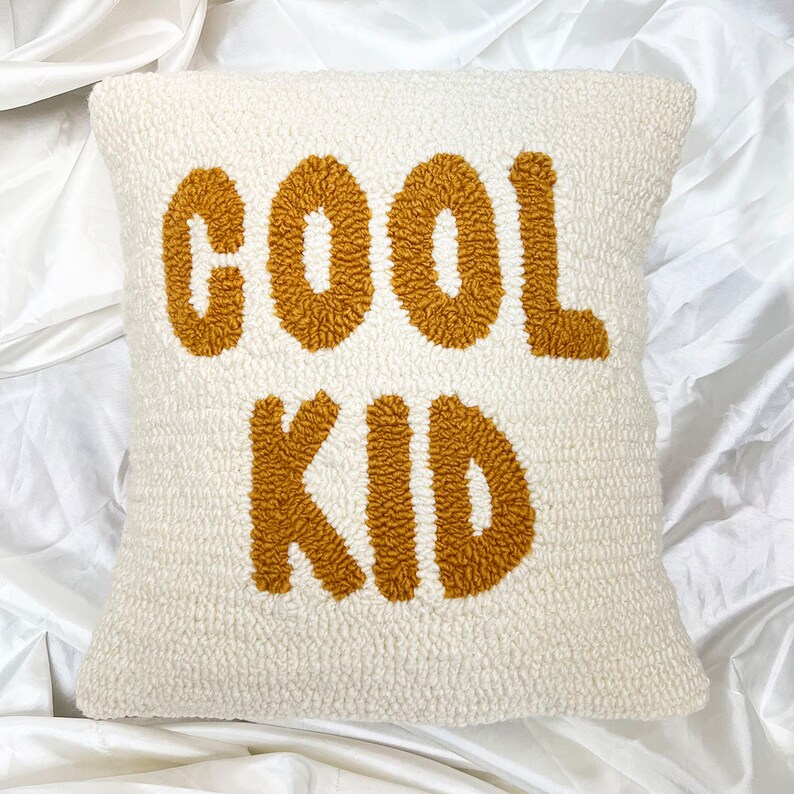 May include: Cream-colored square pillow with the words "COOL KID" embroidered in a textured, golden-brown font. The pillow has a soft, plush texture, and the text is centered on the front.
