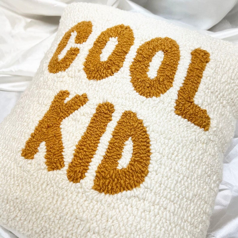 May include: A square, off-white decorative pillow with the words "COOL KID" in a textured, mustard-yellow font. The pillow has a soft, plush appearance, and the text is centered on the front.