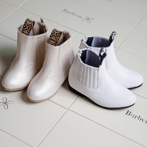 Op de afbeelding: Twee paar enkellaarsjes zijn afgebeeld. Eén paar is parelmoer off-white met stoffen accenten met patronen. Het andere paar is wit met zwarte zolen en gestreepte elastische zijpanelen. De laarzen staan op een lichtgekleurd betegeld oppervlak.