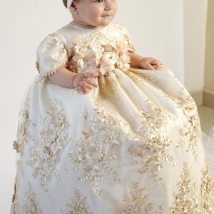 May include: A cream-colored baby dress with intricate gold floral lace detailing. The dress features short sleeves and is adorned with delicate floral embellishments. A white headband with a flower completes the outfit.