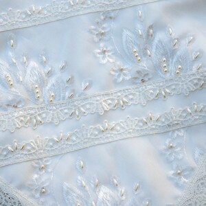 May include: White fabric with delicate lace trim and pearl embellishments. The fabric features embroidered floral designs with pearl accents.