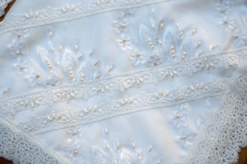 May include: White fabric with delicate lace trim and pearl embellishments. The fabric features embroidered floral designs with pearl accents.