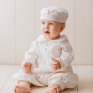Baby is sitting and is wearing our ivory long-sleeved boy's baptism robe, with fine embroidered details, with high-quality fabric to provide comfort to the baby. He has a hat that matches the baptismal robe