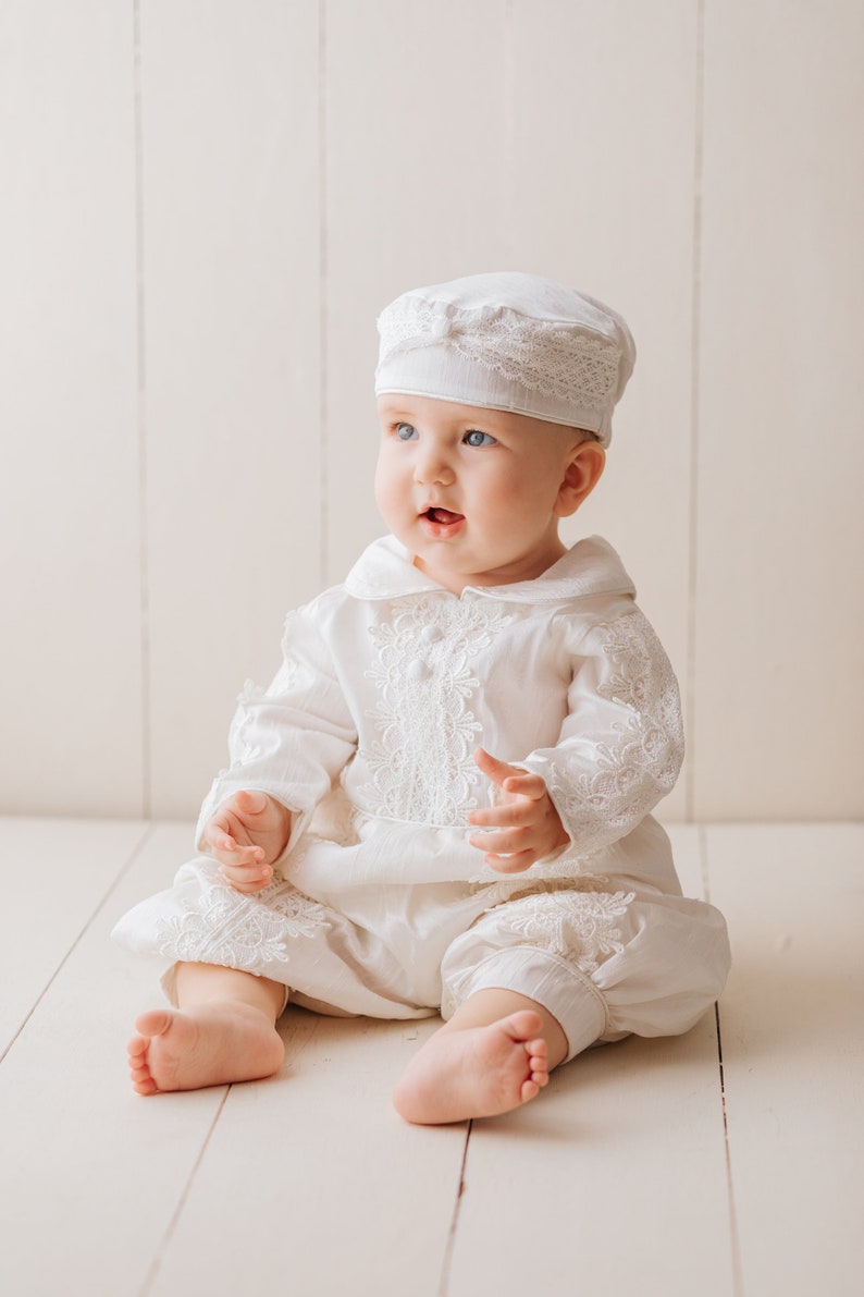 Baby is sitting and is wearing our ivory long-sleeved boy's baptism robe, with fine embroidered details, with high-quality fabric to provide comfort to the baby. He has a hat that matches the baptismal robe