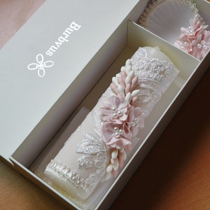 May include: A white box with the word "BULPVUS" printed on the top. The box contains a white candle decorated with lace and pink flowers. The candle is in a white box with a white ribbon.