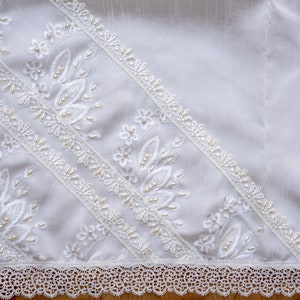May include: White lace fabric with delicate floral embroidery and pearl accents. The fabric has a scalloped edge and is ideal for crafting or decorating.