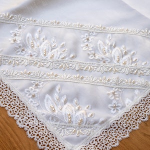 May include: White table runner with delicate lace trim and pearl embellishments. The lace is a delicate pattern with a scalloped edge. The runner is perfect for a wedding or special occasion.