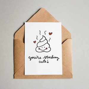 May include: A white card with a cartoon poop emoji with a smiley face and hearts. The text on the card reads "you're stinking cute!"