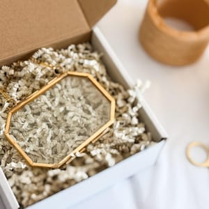 May include: A gold-coloured, geometric picture frame with a glass front sits inside an open white box filled with shredded paper. The frame and packing material are visible. Other decorative items are in the background.