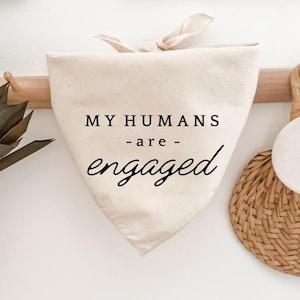 Op de afbeelding: Crèmekleurige hondenbandana met de tekst "MY HUMANS - are - engaged" in zwart schrift. De bandana is bovenaan vastgebonden en hangt aan een houten rek. Een geweven mand en een ronde geweven placemat zijn zichtbaar op de achtergrond.