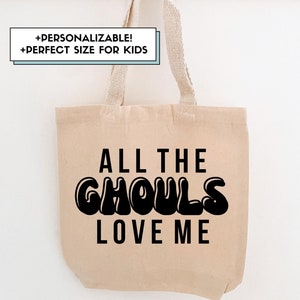 Puede incluir: Una bolsa de lona beige con el texto "ALL THE GHOULS LOVE ME" en letras negras estilo burbuja. La bolsa tiene asas beige a juego y está colgada de un perchero de madera.