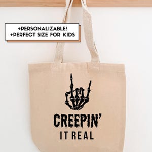 Puede incluir: Una bolsa de lona beige con un gráfico de mano de esqueleto negro haciendo un gesto de rock and roll. El texto "CREEPIN' IT REAL" está debajo del gráfico. La bolsa tiene asas beige y está colgada de un perchero de madera.