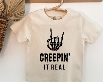 May include: A cream coloured t-shirt with a black skeleton hand making a rock and roll sign. The text "Creepin' It Real" is printed below the hand.