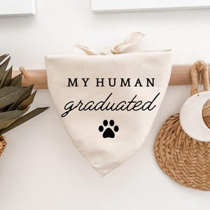 May include: Cream-colored dog bandana with the text "MY HUMAN graduated" in black script, and a black paw print. The bandana is tied at the top and hangs from a wooden rod. A woven basket and a decorative crescent moon are visible.