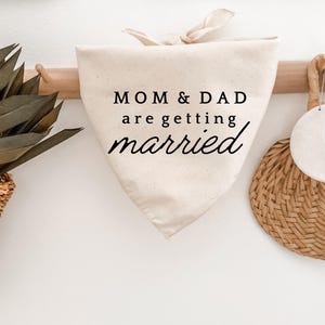 May include: A cream-colored dog bandana with the text "MOM & DAD are getting married" in black script. The bandana is tied at the top and hangs from a wooden rod. A woven basket and a round woven placemat are also visible.