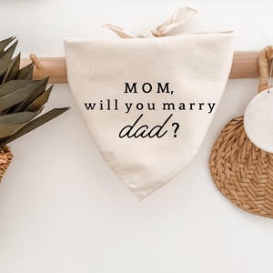 Könnte beinhalten: Cremefarbenes Hundehalstuch mit dem Text "MOM, will you marry dad?" in schwarzer Schrift. Das Halstuch ist oben gebunden und hängt an einer Holzstange. Ein geflochtener Korb und ein dekorativer Gegenstand sind im Hintergrund zu sehen.