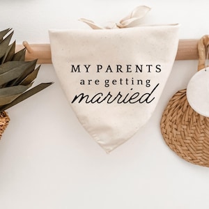 May include: Cream-colored dog bandana with the text "MY PARENTS are getting married" in black script. The bandana is hanging on a wooden rack against a white wall, with decorative items on either side.